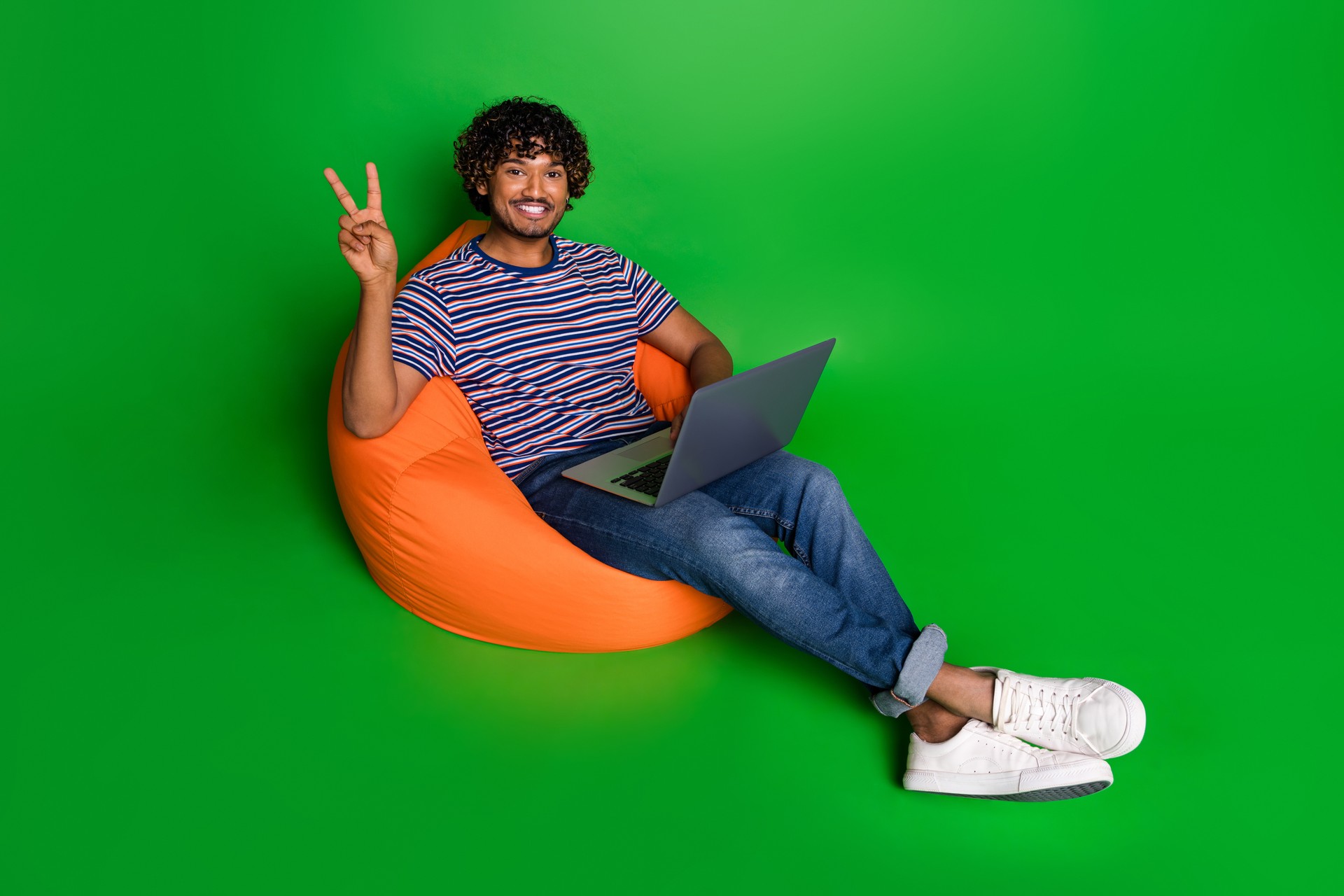 Photo of curly funny man student in t shirt using laptop for work when lying on bean bag show v sign isolated over green color background