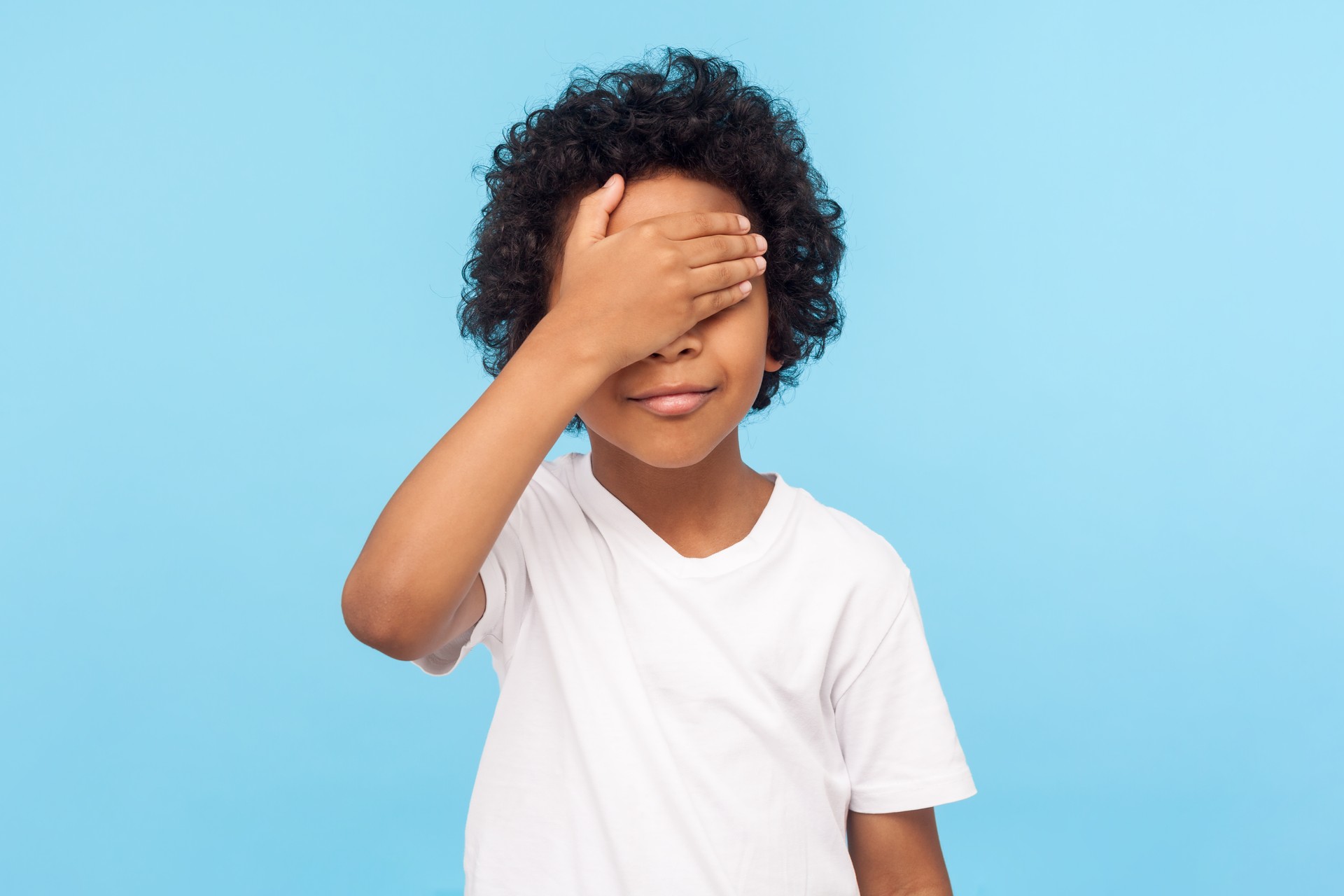 I'm not watching. Portrait of cute little boy with curls covering eyes with hand not to look at something forbidden, hiding eyes