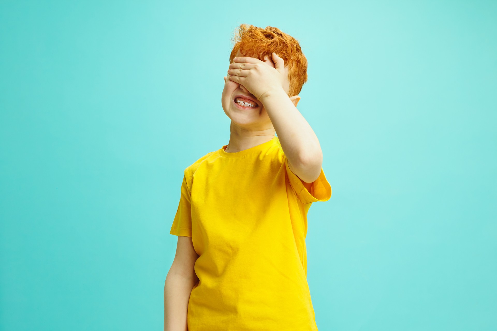 Puzzled worried red haired boy covers eyes with hand, frowns face, being in stupor and bewilderment, doesn't know what to do, isolated over blue background. Child facial expressions concept.